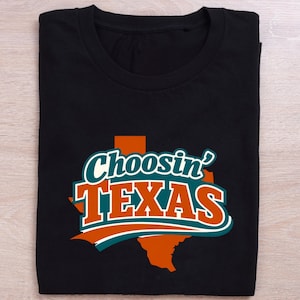 May include: Black t-shirt featuring the phrase "Choosin' TEXAS" in a bold design. The text is white, teal, and orange, set against a Texas state outline. The t-shirt is neatly folded, ready for wear.