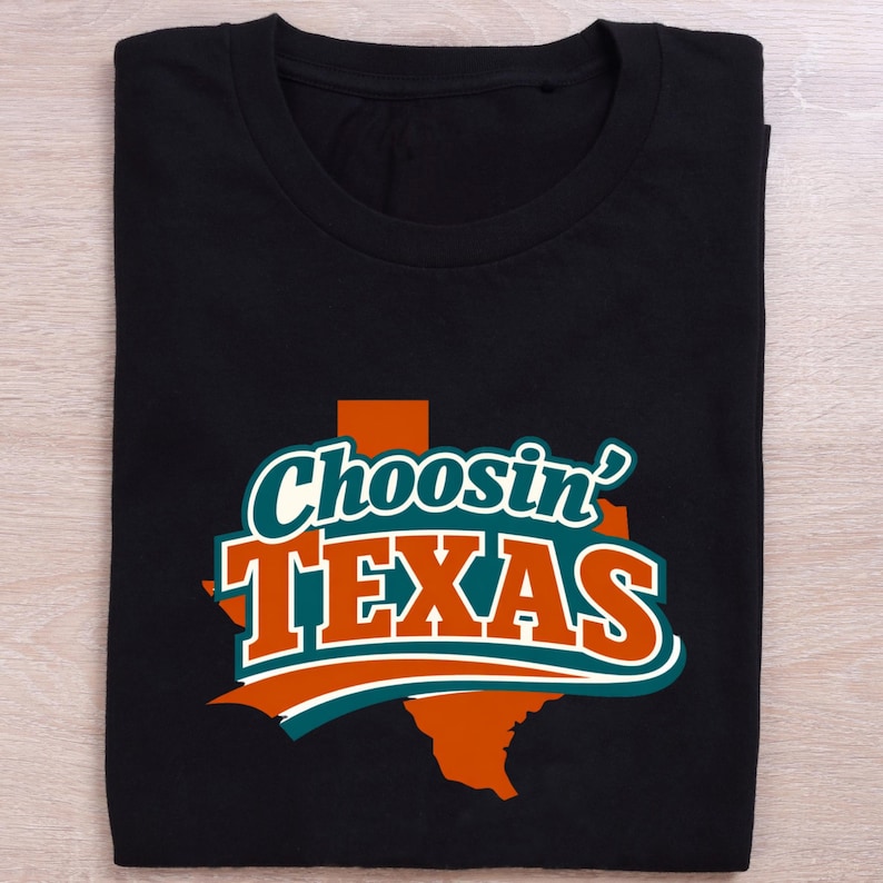 May include: Black t-shirt featuring the phrase "Choosin' TEXAS" in a bold design. The text is white, teal, and orange, set against a Texas state outline. The t-shirt is neatly folded, ready for wear.