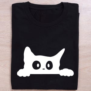 May include: Black t-shirt featuring a white graphic of a cat's face peeking over the bottom edge. The cat has large, round eyes and small paws. The shirt is folded neatly on a light wood surface.