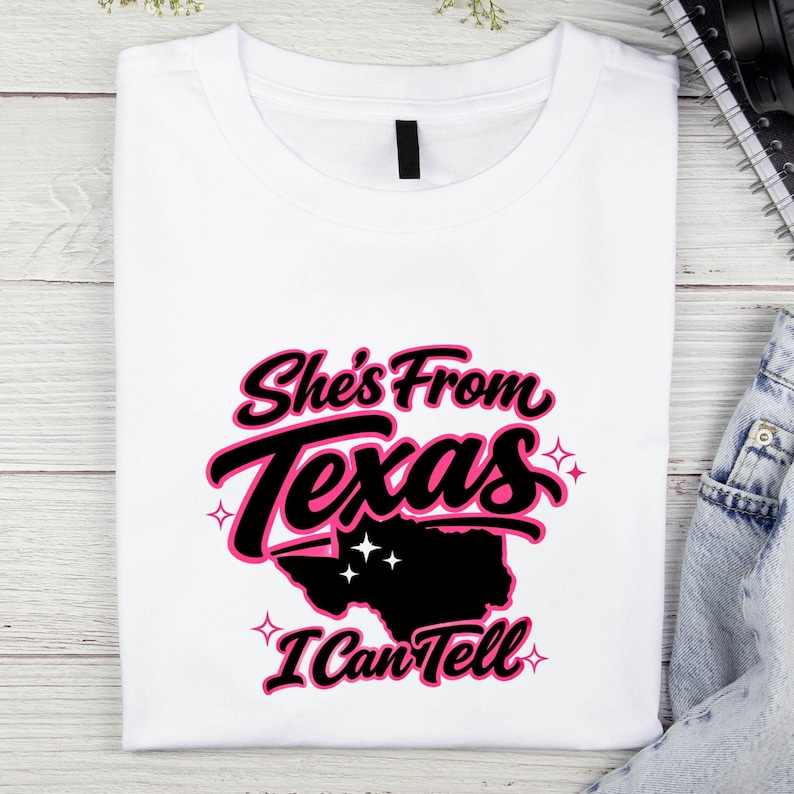 May include: White t-shirt featuring the text "She's From Texas I Can Tell" in black and pink. The design includes a silhouette of the state of Texas. The shirt is folded.