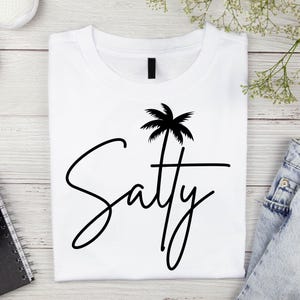 Puede incluir: Camiseta blanca con un gráfico de palmera negra y el texto "Saty" en una fuente cursiva.