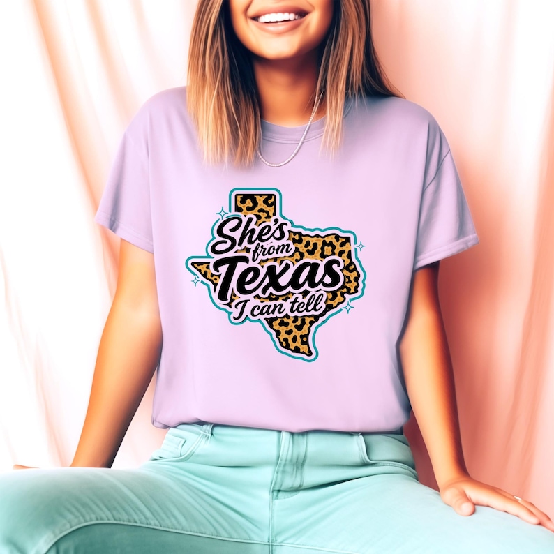 May include: A pale purple t-shirt featuring a graphic of the state of Texas with a leopard print design and the text "She's from Texas I can tell". The design is in black, teal, and yellow.