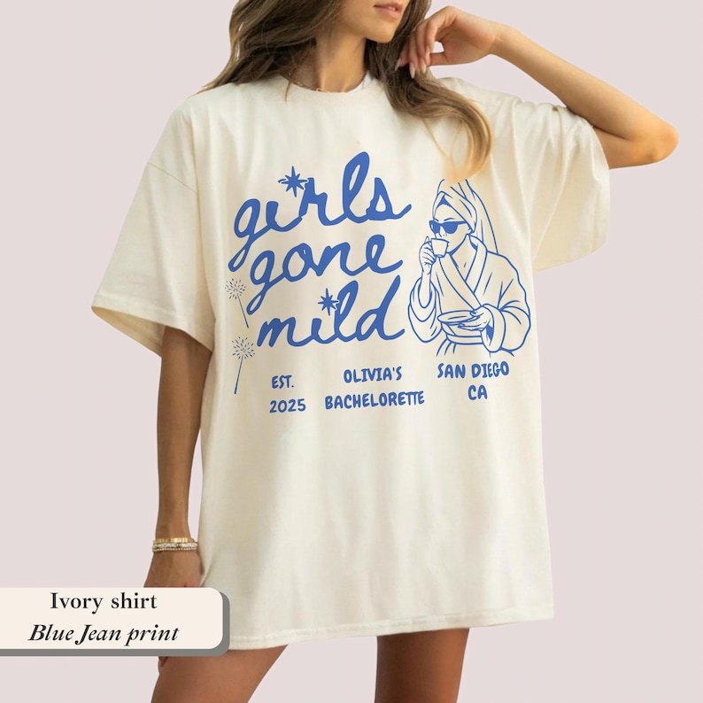 Op de afbeelding: Ivoor t-shirt met een blauwe print met de tekst "girls gone mild" en een tekening van een persoon in een badjas. De tekst bevat ook "Olivia's Bachelorette" en "San Diego, CA".