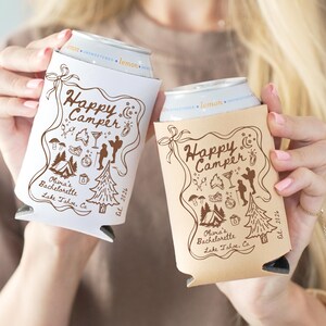 以下が含まれることがあります： キャンプをテーマにした、白とベージュの2つの缶クーラー。クーラーには「Happy Camper」と「Olivia's Bachelorette Lake Tahoe, Ca. Est. 2026」の文字が茶色で書かれています。デザインには、テント、木、その他のキャンプ要素のイラストが含まれています。