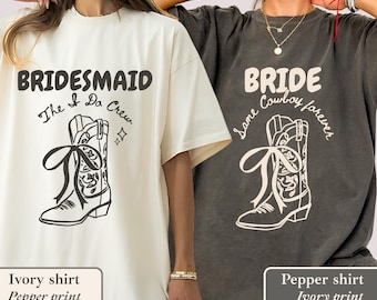 May include: Two t-shirts, one ivory and one pepper, with wedding-themed graphics. The ivory shirt says "BRIDESMAID" above a cowboy boot illustration. The pepper shirt says "BRIDE" above a cowboy boot illustration.
