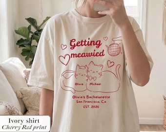 May include: Ivory-colored t-shirt with a cherry red graphic. The design includes the text "Getting meawied" with cat illustrations and the names "Olivia" and "Michael". It also says "Olivia's Bachelorette San Francisco, Ca EST. 2026".