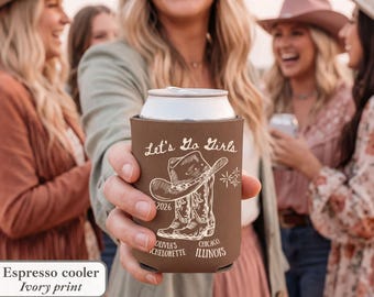 May include: Espresso-colored can cooler with ivory print. The design features a cowboy hat and boots, the text "Let's Go Girls", and "Olivia's Bachelorette Chicago Illinois 2026". The cooler is held by a person in a group of women.