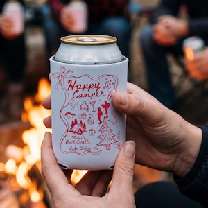 以下が含まれることがあります： 赤いグラフィックと「Happy Camper」の文字、キャンプをテーマにしたイラストが描かれた白い缶クーラー。 「Olivia's Bachelorette Lake Tahoe, CA」の文字も表示されています。 クーラーはキャンプファイヤーの前に保持されています。
