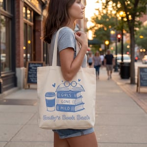 May include: Cream-colored tote bag with a blue graphic of stacked books, a coffee cup, and the text "GIRLS GONE MILD" and "Emily's Book Bach." The bag is carried by a person on a city street.