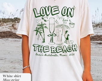 May include: White t-shirt with a green graphic that says "LOVE ON THE BEACH". The design includes palm trees, champagne flutes, and a woman sitting under an umbrella. Text at the bottom reads "Emma's Bachelorette, Miami, 2025".