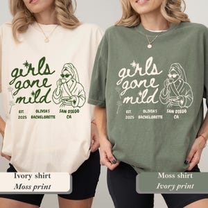 Op de afbeelding: Twee T-shirts, &eacute;&eacute;n ivoorkleurig en &eacute;&eacute;n mosgroen, tonen de tekst "girls gone mild" in een speels lettertype. Het ontwerp bevat een illustratie van een persoon in een badjas en zonnebril, die een kopje vasthoudt. Extra tekst luidt "Est. 2025 Olivia's Bachelorette San Diego CA."