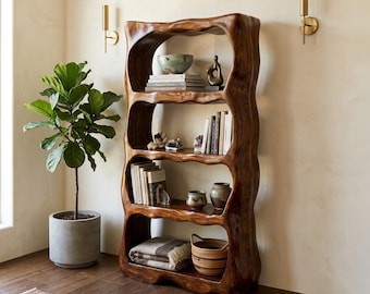May include: A tall, dark brown wooden bookshelf with an organic, wavy frame featuring four open shelves. The shelves are filled with various books, small ceramic bowls, decorative sculptures, a folded textile, and a woven basket.