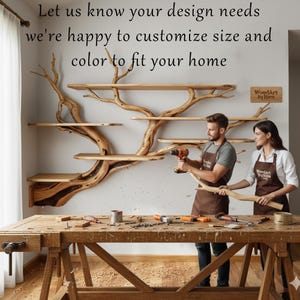 Op de afbeelding: Een unieke houten boomvormige boekenplank met meerdere planken, gemonteerd op een witte muur. De afbeelding toont een man en een vrouw die aan de plank werken. Tekst op de muur zegt: "Laat ons uw ontwerpbehoeften weten, we passen graag de maat en kleur aan uw huis aan."