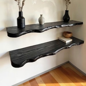 Könnte beinhalten: Zwei schwarze schwebende Holzregale mit naturbelassenen Kanten sind an einer weißen Wand befestigt. Das obere Regal enthält eine schwarze Vase, eine kleine beige Vase und getrocknete Stiele. Das untere Regal zeigt eine Holzschale und ein Buch.