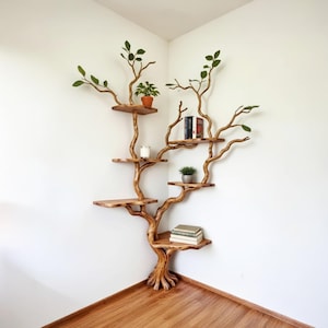 Op de afbeelding: Een houten boomvormige boekenplank met meerdere planken en takken, ontworpen voor een hoek. De planken bevatten kleine potplanten, boeken en een kaars. De boom heeft een natuurlijke houten afwerking en groene bladaccenten.