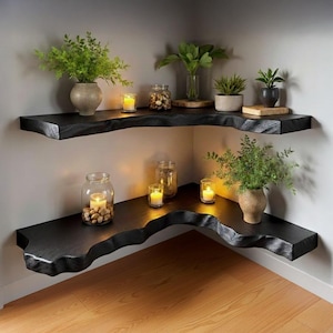 Op de afbeelding: Twee zwarte hoekplanken met een ruwe rand zijn aan een grijze muur bevestigd. De planken tonen potplanten, kaarsen en glazen potten gevuld met decoratieve items. De planken zijn gemaakt van donker hout en hebben een natuurlijke, organische vorm.