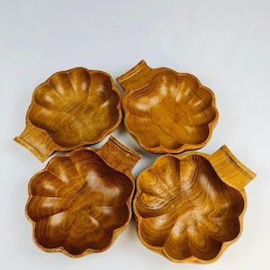 May include: Four wooden serving bowls with a leaf-like design. The bowls are made of a light brown wood with visible grain patterns. Each bowl has a scalloped edge and a small handle. The bowls are arranged on a white surface.