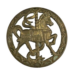 May include: A brass-colored carousel horse in a circular frame. The horse is depicted in a running pose, with a decorative saddle and a pole. The frame has an ornate, repeating pattern around the edge.
