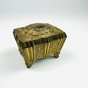 May include: A vintage brass jewelry box with a square shape and ornate floral detailing. The box has a hinged lid with a small, oval-shaped design on top. It rests on four small feet and has a warm, antique gold finish.