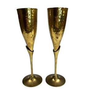 May include: Two vintage champagne flutes with a hammered gold finish. The flutes have a unique design with a slender stem and a wide, textured bowl. The base is round and the overall aesthetic is elegant and sophisticated.