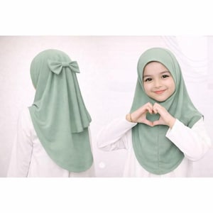 May include: A sage green hijab featuring a rounded front drape and a decorative bow detail at the back. The head covering is shown from both the front and back angles, displaying a smooth, solid-coloured fabric that frames the face and shoulders.