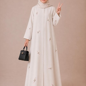 May include: A floor-length beige abaya featuring long sleeves and a front button closure, adorned with small embroidered floral sprigs in green and pink. The garment is paired with a matching beige hijab and a small black structured handbag.