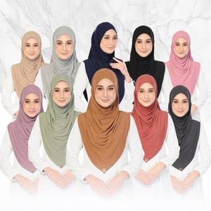 May include: A collection of ten hijabs in various solid colours including beige, grey, navy, black, pink, mauve, sage green, brown, terracotta, and charcoal, each featuring a small metallic logo charm at the bottom centre of the fabric.