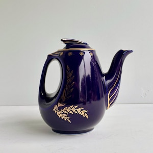 May include: A deep cobalt blue ceramic teapot featuring a glossy finish and gold leaf-style botanical patterns. The teapot has a curved handle, a pointed spout, and a matching lid with gold trim accents along the rim and the base of the handle.