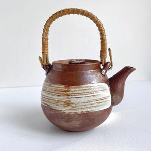 May include: A round, brown ceramic teapot featuring a textured, horizontal white brushstroke pattern across the center. The teapot includes a matching lid and a curved, light brown woven rattan handle attached to two small loops on the top rim.