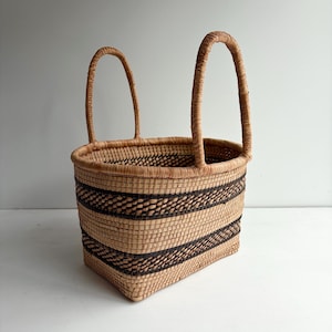 May include: A rectangular woven basket featuring two tall, upright handles. The basket is constructed from light tan natural fibers and is accented with two horizontal bands of dark brown woven patterns encircling the main body.