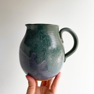 May include: A ceramic pitcher with a rounded body and a curved handle. The exterior features a mottled glaze with deep forest green tones transitioning into a muted purple hue at the base. The rim has a small spout for pouring liquids.