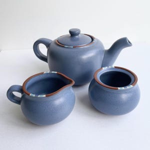 May include: A blue ceramic tea set with a teapot, creamer, and sugar bowl. The set has a speckled finish with brown trim and a decorative band of blue and white stripes. The teapot has a lid and a curved handle.