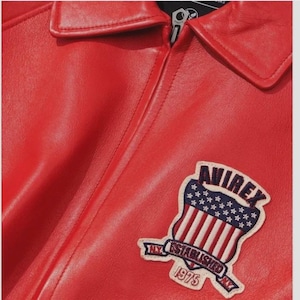 May include: Close-up of a red leather jacket with a collar and zipper. A patch on the jacket features a shield design with the text "AVIREX NY ESTABLISHED 1975". The jacket has a smooth, glossy finish.