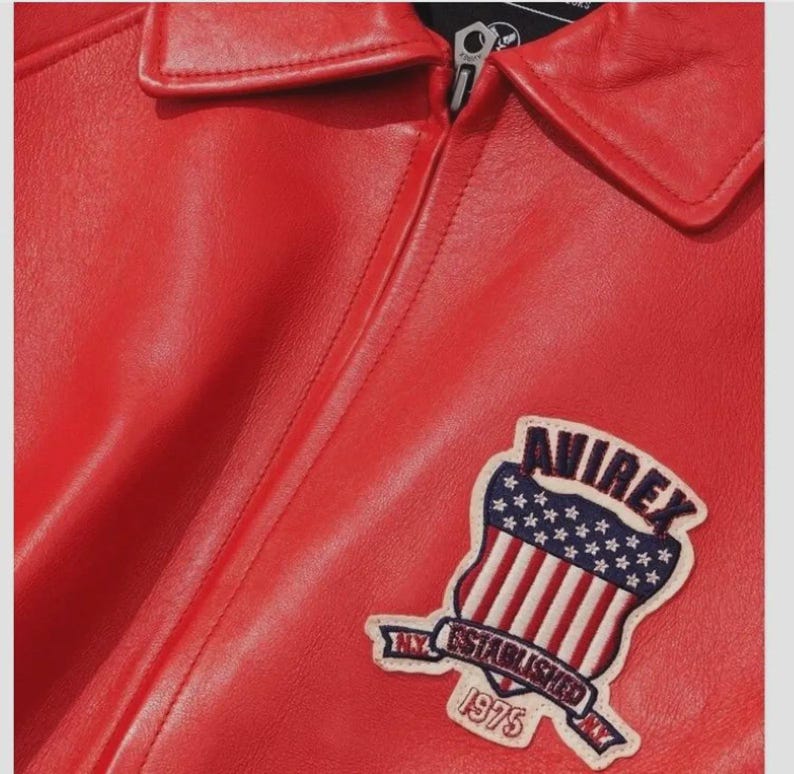 May include: Close-up of a red leather jacket with a collar and zipper. A patch on the jacket features a shield design with the text "AVIREX NY ESTABLISHED 1975". The jacket has a smooth, glossy finish.