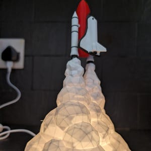 May include: A white and red space shuttle lamp with a geometric cloud base. The lamp is plugged into a wall outlet. The shuttle is depicted taking off, with white smoke plumes. The lamp is lit, casting shadows.