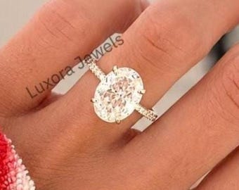 May include: A gold ring featuring a large oval-cut center stone held by four prongs, with a thin band accented by small pavé-set stones. The text Luxora Jewels is visible near the ring, which is worn on a finger against a blurred background.