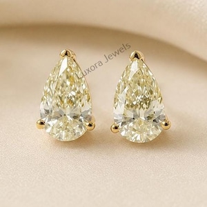 May include: A pair of pear-shaped stud earrings featuring faceted, light yellow stones set in yellow gold prongs. The earrings are positioned side-by-side, showcasing the brilliant cut of the stones and the metallic lustre of the gold settings.