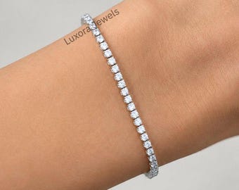 May include: A silver-toned tennis bracelet featuring a continuous row of round, clear, faceted stones set in a metallic frame. The bracelet is marked with the text Luxora Jewels in two locations along the length of the piece.