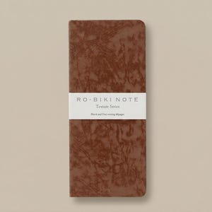 May include: A rectangular notebook with a dark brown, marbled texture cover. A white horizontal paper band wraps around the centre, featuring the text RO-BIKI NOTE, Texture Series, and Blank and Free writing 60pages in black lettering.