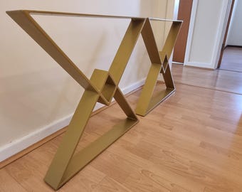 May include: Two gold-colored metal table legs with an X-shaped design. The legs are angled and have a geometric aesthetic. The legs are made of metal and are designed to support a tabletop. The legs are shown on a wooden floor.