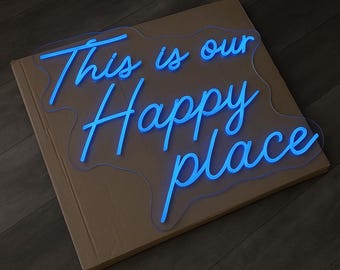 May include: A blue neon sign with the words "This is our Happy place" in a cursive font. The sign is mounted on a cardboard surface, and the background is a dark wood floor.