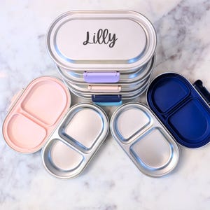 May include: Silver metal lunchboxes, stacked with the name "Lilly" in black script on the top. Open lunchboxes in pink and blue show divided compartments. The lunchboxes have colourful clasps. Ideal for packed lunches.