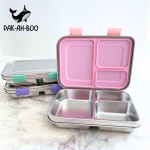 Op de afbeelding: Roestvrijstalen lunchboxen met roze, mintgroene en paarse accenten. De open lunchbox onthult een roze interieur met meerdere compartimenten. Het logo "PAK-AH-BOO" is zichtbaar in de linkerbovenhoek, met een walvis met een feesthoed.