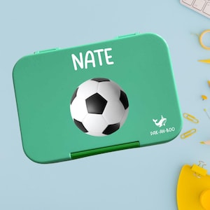 Puede incluir: Una fiambrera verde con un balón de fútbol y el nombre "NATE" en blanco. La fiambrera también presenta un pequeño logotipo de ballena y el texto "PAK-AH-BOO". La fiambrera es rectangular con esquinas redondeadas.