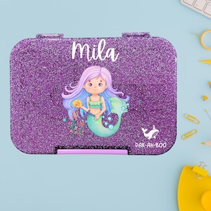 Könnte beinhalten: Eine lila Glitzer-Lunchbox mit einer Meerjungfrau-Illustration und dem Namen "Mila" in weißer Schrift. Die Meerjungfrau hat blau-violettes Haar, ein grünes Oberteil und einen türkisfarbenen Schwanz. Die Lunchbox hat einen schwarzen Verschluss und die Worte "PAK-AH-BOO".