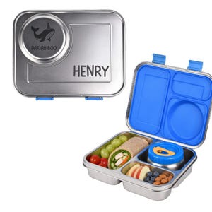 Puede incluir: Una fiambrera bento rectangular de acero inoxidable con un divisor interior de silicona azul. La tapa presenta el nombre grabado HENRY y un gráfico de ballena con el texto PAK-AH-BOO. La caja abierta contiene un wrap, uvas, tomates y frutos secos.