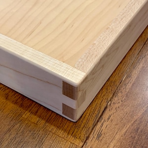 May include: Close-up of a wooden box with dovetail joints. The box is made of light-colored wood, with a smooth, natural finish. The dovetail joints are visible at the corners, showcasing the craftsmanship. The box sits on a wooden surface.