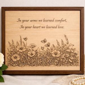 May include: Wooden plaque with a dark brown frame. The light wood surface features engraved text: "In your arms we learned comfort, In your heart we learned love." Below the text is an engraved floral and butterfly design.