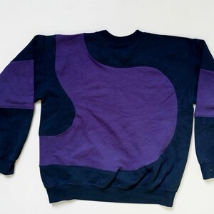 Puede incluir: Una sudadera azul marino con un gran dise&ntilde;o abstracto p&uacute;rpura en la parte delantera y las mangas. La sudadera tiene cuello redondo y mangas largas. El dise&ntilde;o es una mezcla de colores y formas s&oacute;lidas.