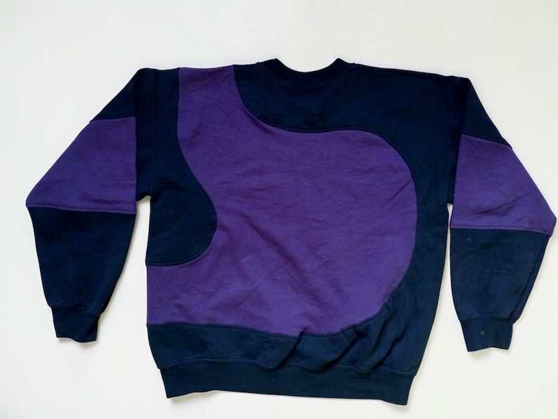 Puede incluir: Una sudadera azul marino con un gran dise&ntilde;o abstracto p&uacute;rpura en la parte delantera y las mangas. La sudadera tiene cuello redondo y mangas largas. El dise&ntilde;o es una mezcla de colores y formas s&oacute;lidas.